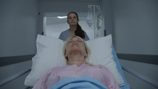 老年女性患者术后在有呼吸管的绞痛床上睡觉 年轻的护士把老年妇女运送到医院走廊下面的病房 在医疗中心工作的医务人员 — 图库视频影像