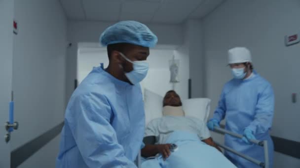 医師や救急隊員はアフリカ系アメリカ人の患者と手術室にストレッチャーを押す ネックブレースを持つ男はガーニーに横たわっている 複数の民族が緊急部門で働いている フロントビュー トラッキングショット — ストック動画