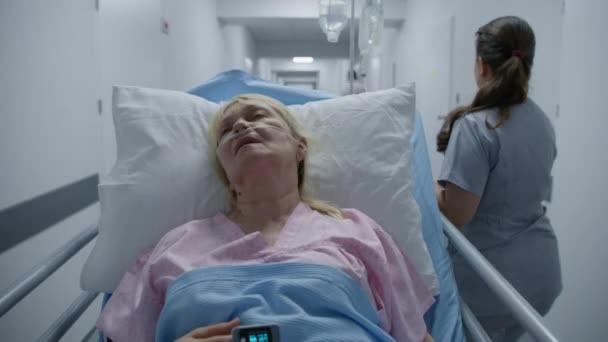 年轻的护士把老年妇女从医疗中心的走廊运送到医院病房 老年病人躺在带有呼吸管 脉动血氧计和滴注的骨科上 在现代诊所工作的医务人员 — 图库视频影像