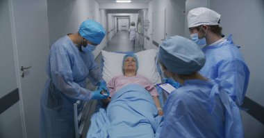 Doktorlar, hemşireler ve sağlık görevlileri hasta yaşlı kadını tıbbi tesis koridorunda muayene ediyorlar. Doktor, nabız oksimetresini kontrol ediyor. Sağlık personeli acil serviste çalışıyor. Yakınlaştır.