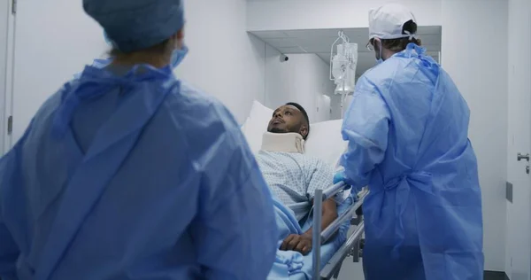 医師や救急隊員は 患者と手術室にストレッチャーを押します ネックブレースを持つアフリカ系アメリカ人男性は ガーニーに横たわっています 複数の民族がクリニックで働いている スローモーション バックビュー トラッキングショット — ストック写真