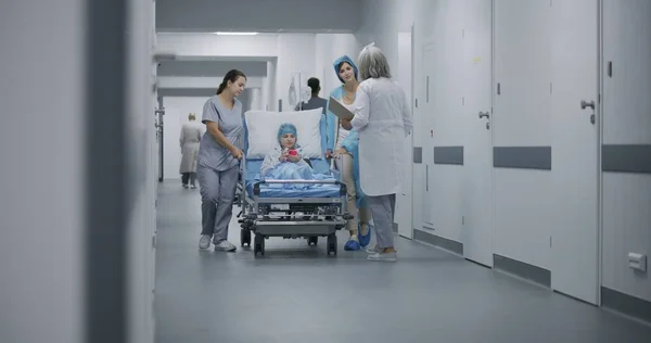 看護師と子供の母親は 診療所の廊下で若い女の子とガーニーを押しました 女性医師はストレッチャーの子供に横たわり 手術前に彼女と話をする 救急部に勤務する医療従事者 — ストック写真