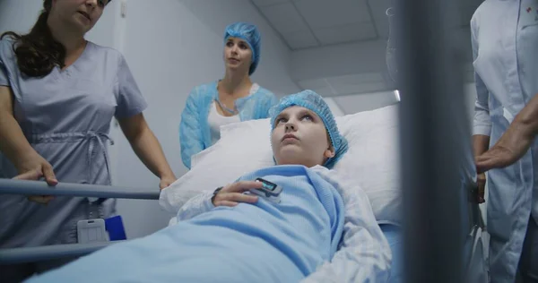 医学は 医療センターの廊下の手術後に患者を病院室に搬送する ガーニーに横たわる若い女の子は 彼女の指にパルスオキシメートルを見て 母親と話しています 職場の医療スタッフ — ストック写真