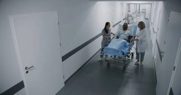女性医師と看護師がアフリカ系アメリカ人の患者と病院の廊下に立っている 専門の医師は手術室に男性を運ぶ セキュリティカメラビュー サーベイランスシステム — ストック写真