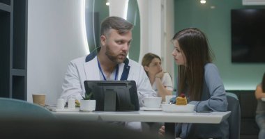 Profesyonel doktor hastane kafesindeki dijital tablet bilgisayardaki hastaya tıbbi testlerin sonuçlarını gösteriyor ve bunu tartışıyor. Kadın doktor arka planda yemek yiyor. Hastane kantininde tıbbi personel.