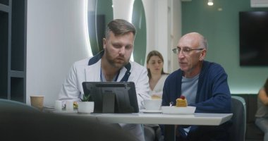 Yetişkin doktor kıdemli hastayla konuşur ve MRI veya CT taramasının dijital tablet bilgisayardaki sonuçlarını açıklar. Kadın doktor dinleniyor ve arka planda telefonunu kullanıyor. Hastane kantininde tıbbi personel.