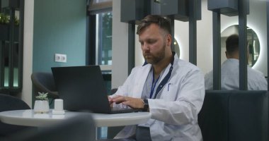 Erkek doktor modern klinik kafesinde oturuyor. Sağlık uzmanı dizüstü bilgisayarda çalışır, klavyede yazı yazar ve hastayla tedavi hakkında sohbet eder. Tıp merkezi kafeteryasında tıbbi personel. Yavaş çekim.