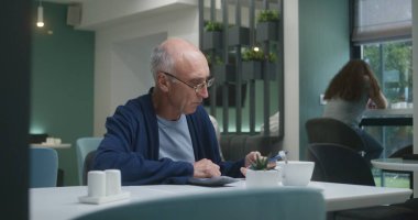 Yaşlı hasta hastane kafeteryasında cep telefonu kullanıyor. Garson kıdemli adama yemek getiriyor. Yetişkin doktor arka planda yemek yiyor. Tıbbi personel ve hastalar tıp merkezi kantininde dinleniyor..