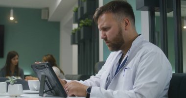 Profesyonel doktor hastasının test sonuçlarını dijital tablet bilgisayarda izler ve kahve içer. Kadın doktor arka plandaki kadınla konuşuyor. Modern hastane kantininde çalışan tıbbi personel.