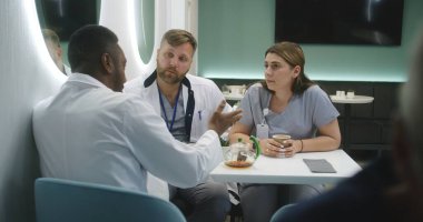 Afrikalı Amerikalı doktor klinikteki kafede iş arkadaşlarıyla oturuyor. Profesyonel sıhhiyeciler yemek boyunca konuşur. MRI beyin taraması görüntülü dijital tablet. Hastane veya tıp merkezi kafeteryasında tıbbi personel.