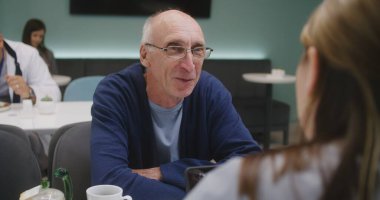 Yaşlı hasta kadın doktorla klinikte oturuyor. Sağlık uzmanı, tıbbi tanı sonuçlarını adamla tartışıyor. Sıhhiyeci arka planda yemek yiyor. Hastane ya da Tıp Merkezi Kafeteryası.