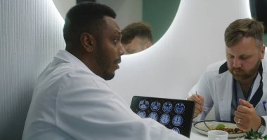 Afrikalı Amerikalı doktor klinikteki kafede iş arkadaşlarıyla oturuyor. Sağlık uzmanları öğle yemeğinde konuşurlar. MRI beyin taraması görüntülü dijital tablet. Hastane veya tıp merkezi kantininde tıbbi personel.