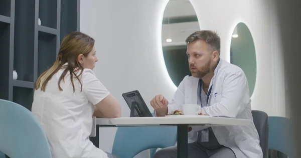 Medic sits in hospital cafeteria with his colleague. Adult doctor eats dinner, examines test results of patient on digital tablet computer and talks to nurse. Medical staff have break in clinic cafe.