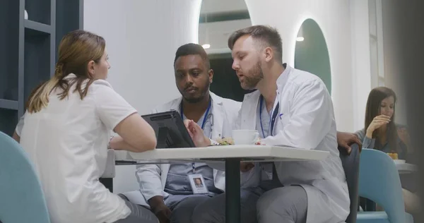 Multiethnic medics sit in clinic cafeteria. Caucasian doctor eats dinner, uses digital tablet, shows tests results to his colleagues. Medical staff discuss work during break in modern hospital cafe.