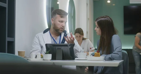 Professional doctor shows medical tests results to patient on digital tablet computer in hospital cafe and discusses it. Female medic eats dinner in the background. Medical staff in hospital canteen.