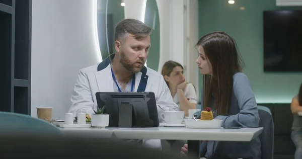 Professional doctor shows medical tests results to patient on digital tablet computer in hospital cafe and discusses it. Female medic eats dinner in the background. Medical staff in hospital canteen.