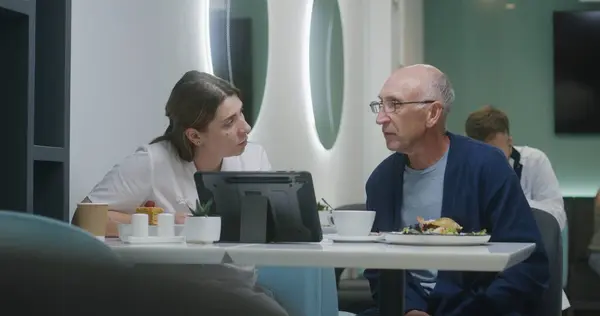 Female doctor shows medical tests results on digital tablet computer in clinic cafeteria and discusses it with elderly man. Adult medic eats meal in background. Medical staff rest in hospital canteen.