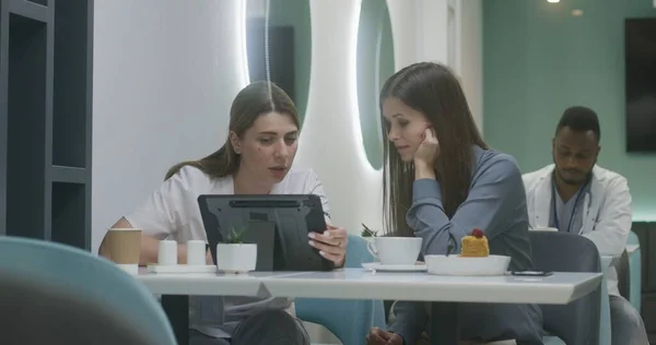 Professional female doctor shows medical tests results to patient on digital tablet computer in clinic cafe and discusses it. Adult medic eats dinner in background. Medical staff in hospital canteen.