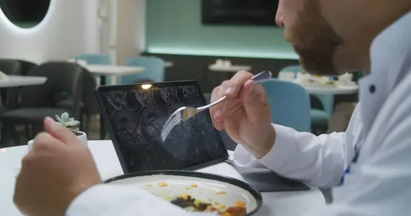 Professional doctor watches image of MRI or CT scan using digital tablet in clinic cafe. Medic eats his dinner, examines brain scanning results of patient. Medical staff have meal in hospital canteen.