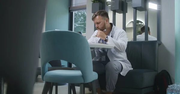 Professional doctor uses digital tablet in modern clinic cafe during break. Medic eats dinner, watches medical tests results of patient or surfs the Internet. Medical staff in hospital dining room.