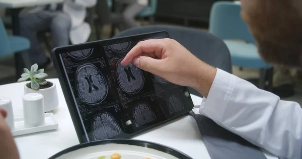 Doctor watches image of MRI or CT scan using digital tablet in hospital cafe. Medic eats his dinner, analyzes brain scanning results of patient. Medical staff have meal in clinic cafeteria. Close up.