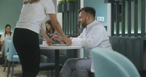 Adult doctor works on digital tablet and eats his dinner in hospital cafe. Cafeteria worker brings meal for medic. Patient uses her phone in the background. Medical staff have break in clinic canteen.