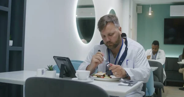 Adult doctor eats dinner and uses digital tablet computer in hospital cafeteria, another medic sits down with test results. Doctor talks with colleague in clinic cafe. Medical staff have break.