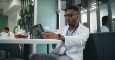 Afrikalı Amerikalı doktor tatilde modern klinik kafesinde dijital tablet kullanır. Sıhhiye çay içer, hastaların test sonuçlarını izler ya da internette sörf yapar. Hastane kantininde tıbbi personel.