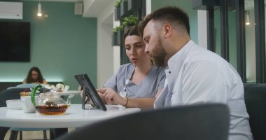 Kadın doktor, erkek meslektaşıyla birlikte modern klinik kafesinde oturuyor. Dijital tablet kullanıyor. Profesyonel doktorlar konuşur, tıbbi testlerin sonuçlarını inceler, teşhisi tartışırlar. Hastane kantininde tıbbi personel çalışıyor..