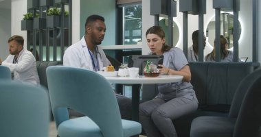 Kadın doktor, Afrikalı Amerikalı meslektaşıyla modern klinikte oturuyor, dijital tablet kullanıyor. Profesyonel sağlık görevlileri konuşur, tıbbi test sonuçlarını izler, iş tartışır. Hastane kantininde tıbbi personel.