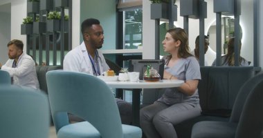 Kadın doktor, Afrikalı Amerikalı meslektaşıyla modern klinikte oturuyor, dijital tablet kullanıyor. Profesyonel sağlık görevlileri konuşur, tıbbi test sonuçlarını izler, iş tartışır. Hastane kantininde tıbbi personel.