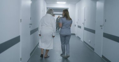 Dijital tabletli kadın doktor, yaşlı bir kadınla klinik koridorunda yürüyen hemşire, hastaların prosedürlerden sonra hastane koğuşuna gitmesine yardım ediyor. Sağlık personeli, hastane koridorundaki hastalar..