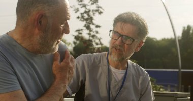 Doktorun hastasıyla bankta otururken ve omzunu muayene ederken yakın çekim. Yaşlı adam ağrı şikâyetini sağlık uzmanına iletiyor. Sıhhiye müşteriye danışıyor ve tedavisini dışarıda tartışıyor.