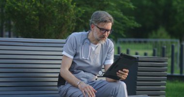 Üniformalı doktor modern hastanenin dışında bankta oturur, dijital tablet kullanılarak online randevu sırasında tedavi hakkında hastalarla konuşur. Klinik ya da Tıp Merkezi 'nin tıbbi personeli. Sağlık hizmetleri sistemi.