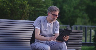 Üniformalı doktor modern hastanenin dışında bankta oturur, dijital tablet kullanılarak online randevu sırasında tedavi hakkında hastalarla konuşur. Klinik ya da Tıp Merkezi 'nin tıbbi personeli. Sağlık hizmetleri sistemi.