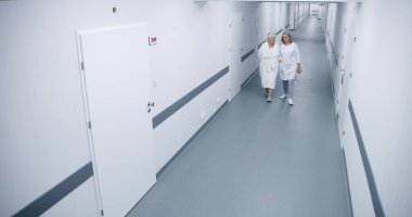 Parlak klinik koridoru: Kadın doktor ve yaşlı kadın hastane koğuşuna doğru yürüyorlar, tedavi hakkında konuşuyorlar. Modern tıbbi tesiste tıbbi personel çalışıyor. Güvenlik kamerası görüntüsü. Yüksek açı.