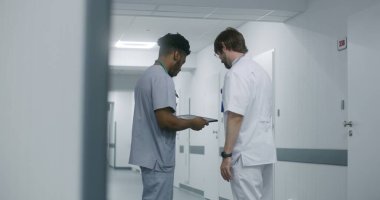 Afro-Amerikalı doktor, meslektaşı ile sağlık merkezi koridorunun ortasında duruyor. Genç doktor diğer tıbbi hasta tedavisi ile görüşüyor ve dijital tablet bilgisayarda test sonuçlarını gösteriyor.