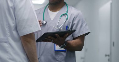 Afro-Amerikan sıhhiyeci dijital tablet bilgisayarda tıbbi test sonuçlarını başka bir doktora gösteriyor. Çok kültürlü doktorlar hastane koridorunun ortasında tedavi yöntemlerini tartışırlar. Tıbbi personel iş başında..