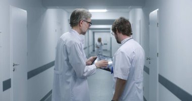 Olgun doktor MRI ve CT beyin taraması görüntülerini dijital tablet kullanarak meslektaşına gösteriyor. Profesyonel sıhhiyeciler ayakta durup klinik koridorunda yürürler, iş tartışırlar. Hastane personeli, hastalar koridorda..