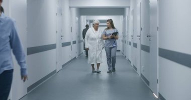 Kadın sıhhiyeci, dijital tabletli hemşire ve yaşlı kadın sohbeti, prosedürlerden sonra klinik koridoru boyunca hastane odasına yürüyor. Sağlık personeli, doktorlar, hastane koridorundaki hastalar..