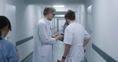 Olgun doktor MRI ve CT beyin taraması görüntülerini dijital tablet kullanarak meslektaşına gösteriyor. Profesyonel sıhhiyeciler ayakta durup klinik koridorunda yürürler, iş tartışırlar. Hastane personeli, hastalar koridorda..