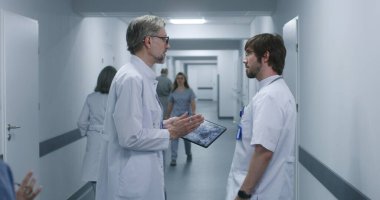 Olgun doktor MRI ve CT beyin taraması görüntülerini dijital tablet kullanarak meslektaşına gösteriyor. Profesyonel sıhhiyeciler ayakta durup klinik koridorunda yürürler, iş tartışırlar. Hastane personeli, hastalar koridorda..