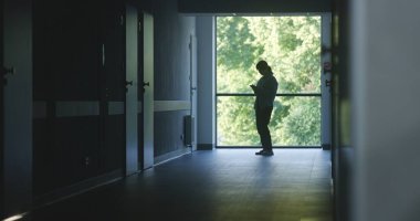 Klinik koridoru: Doktorlar ve profesyonel sağlık görevlileri yürüyor. Dijital tabletli hemşire pencerenin yanında duran kadın hastaya geliyor. Karanlık hastane veya tıp merkezi koridorundaki tıbbi personel ve hastalar.