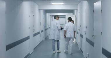 Sıhhiyeciler klinik koridorunda yürürler, iş tartışırlar. Olgun doktor MRI ve CT beyin taraması görüntülerini dijital tablet kullanarak meslektaşına gösteriyor. Hastane ya da tıp merkezi koridorundaki hasta ve sağlık personeli.