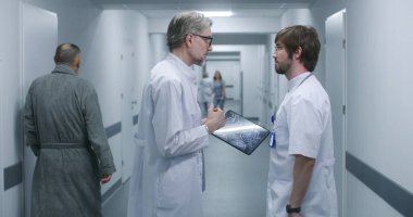 Olgun doktor MRI ve CT beyin taraması görüntülerini dijital tablet kullanarak meslektaşına gösteriyor. Profesyonel sıhhiyeciler ayakta durup klinik koridorunda yürürler, iş tartışırlar. Hastane personeli, hastalar koridorda..