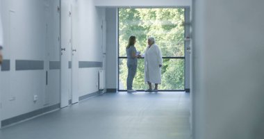 Hastane koridoru: Doktorlar ve profesyonel sağlık görevlileri yürüyor. Dijital tabletli hemşire pencerenin yanında duran yaşlı kadın hastaya geliyor. Klinikteki veya tıp merkezi koridorundaki tıbbi personel ve hastalar.
