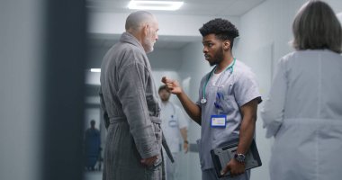 Afrikalı Amerikalı doktor, klinik koridorunda sağlığıyla ilgili eski bir hastaya danışıyor. Kıdemli adam profesyonel doktorla konuşuyor. Hemşire tekerlekli sandalyesini hastane koridorundan revire taşıyor..