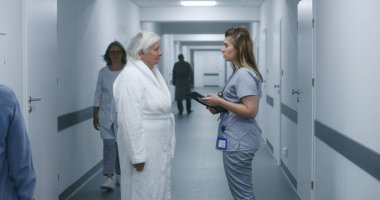 Kadın doktor, dijital tabletli hemşire ve hasta sohbeti. Doktor, prosedürlerden sonra yaşlı bir kadınla modern klinik koridorunda duruyor. Hastane ya da tıp merkezi koridorundaki hasta ve sağlık personeli.