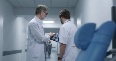 Olgun doktor MRI ve CT beyin taraması görüntülerini dijital tablet kullanarak meslektaşına gösteriyor. Profesyonel sıhhiyeciler ayakta durup klinik koridorunda yürürler, iş tartışırlar. Hastane personeli, hastalar koridorda..