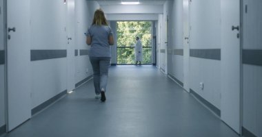 Klinik koridoru: Doktorlar ve profesyonel sağlık görevlileri yürüyor. Dijital tabletli hemşire pencerenin yanında duran yaşlı kadına geliyor. Modern hastane veya tıp merkezi koridorundaki tıbbi personel ve hastalar.